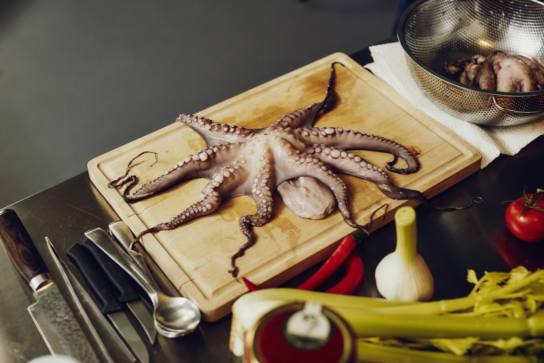 Raw octopus on a wooden cutting board surrounded by fresh vegetables and cooking implements.