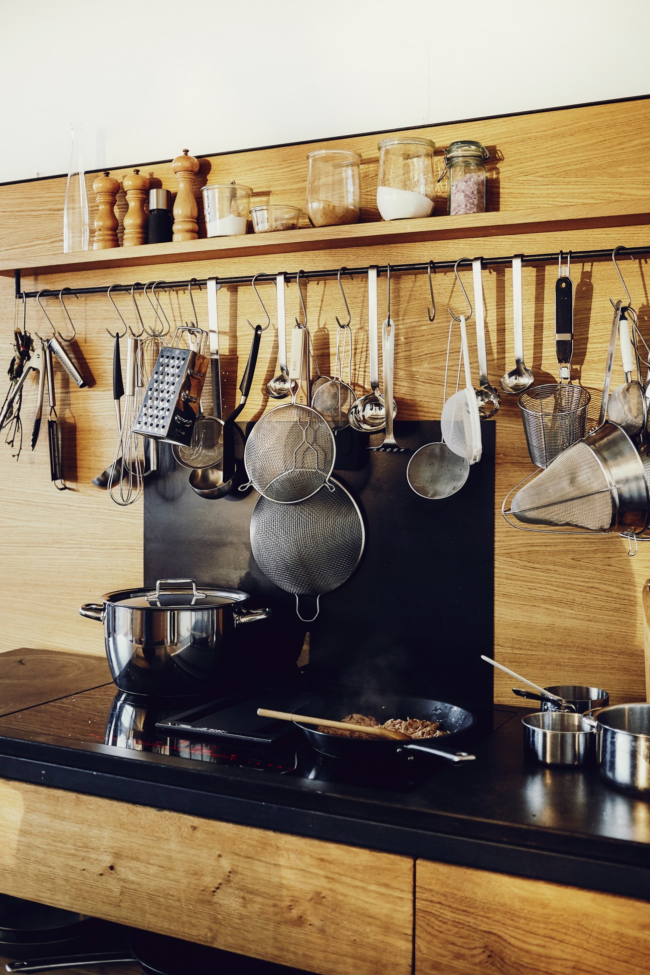 Organized kitchen wall with wooden shelving displaying cooking utensils, pans, and ingredients in jars.