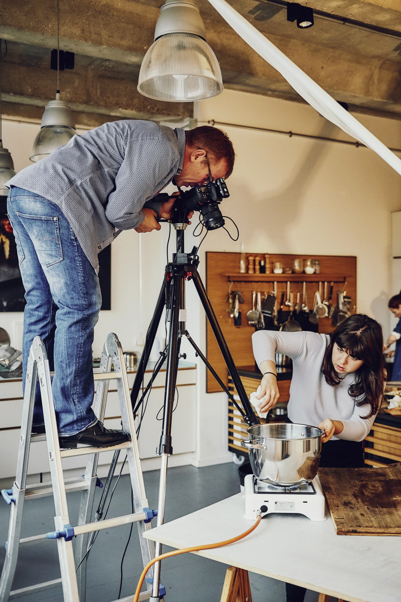 Food photographer on ladder shooting a pot on stovetop with female assistant below in studio kitchen.