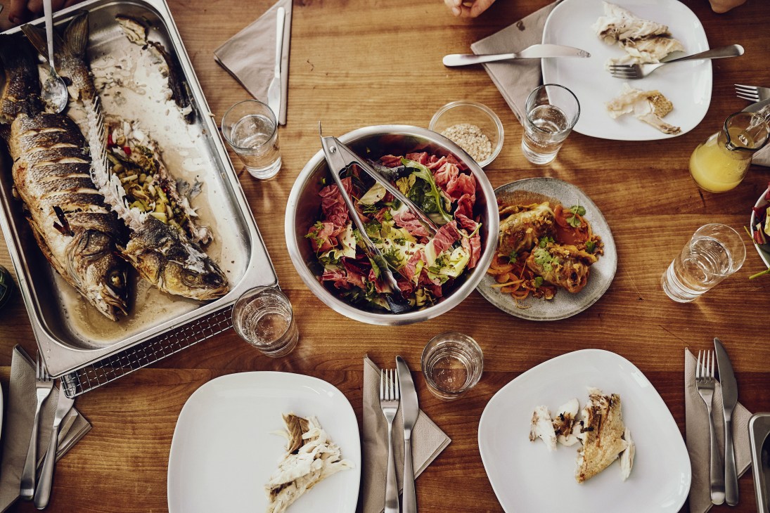 Overhead view of a wooden table set with roasted sea bass, salad, white plates, glasses of water, and cutlery.