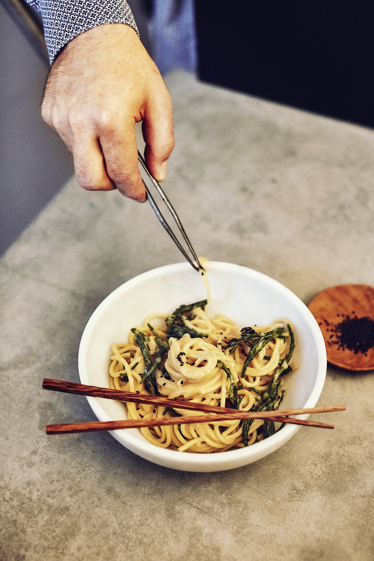 A hand using tweezers to adjust noodles in a white bowl of food on a concrete surface.