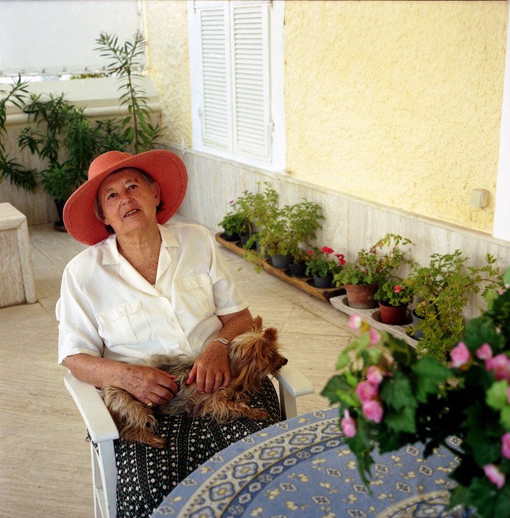 Daphne Wookey sits in a yellow-walled courtyard wearing a red hat and white shirt, holding a small dog, surrounded by potted flowers.