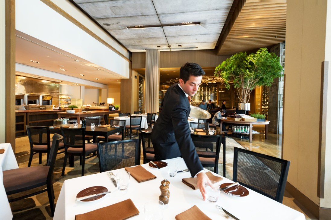 A maître d' in dark suit stands attentively in an upscale restaurant dining room with modern furnishings and warm lighting.