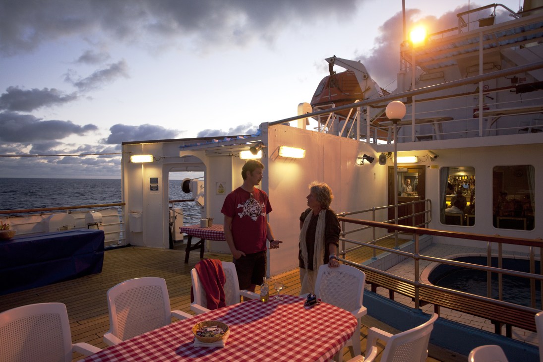 Two passengers stand on the deck of RMS St Helena at sunset, with ship structures and calm ocean visible behind them.
