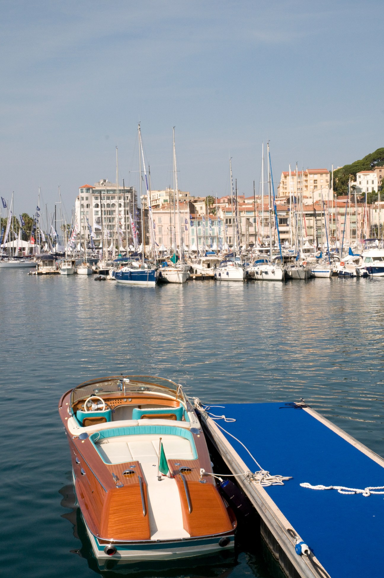 A classic wooden runabout with turquoise and brown trim moored in Cannes harbor with sailing yachts and waterfront buildings in the background.