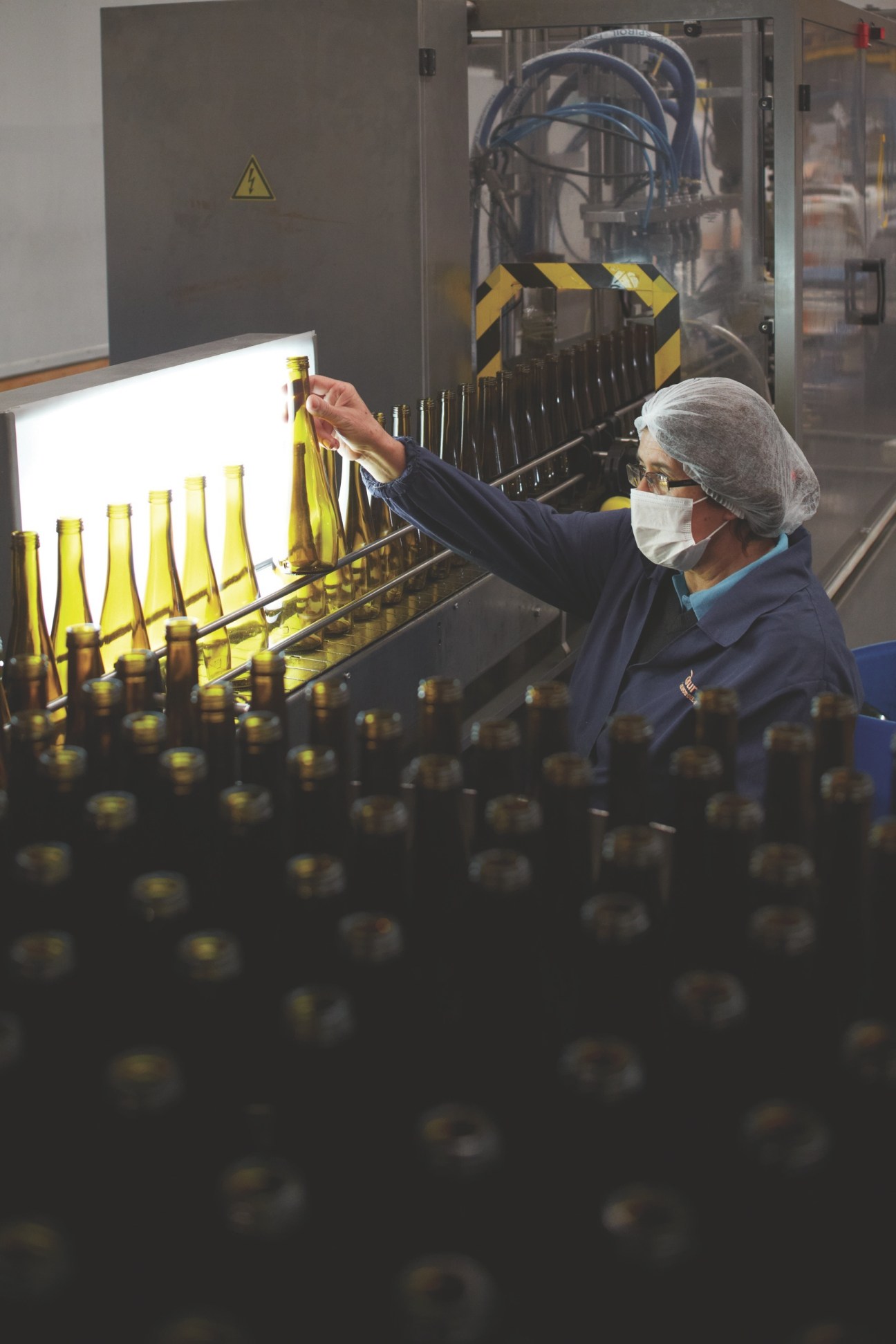 Factory worker in hairnet and mask inspecting yellow-capped olive oil bottles on production line.