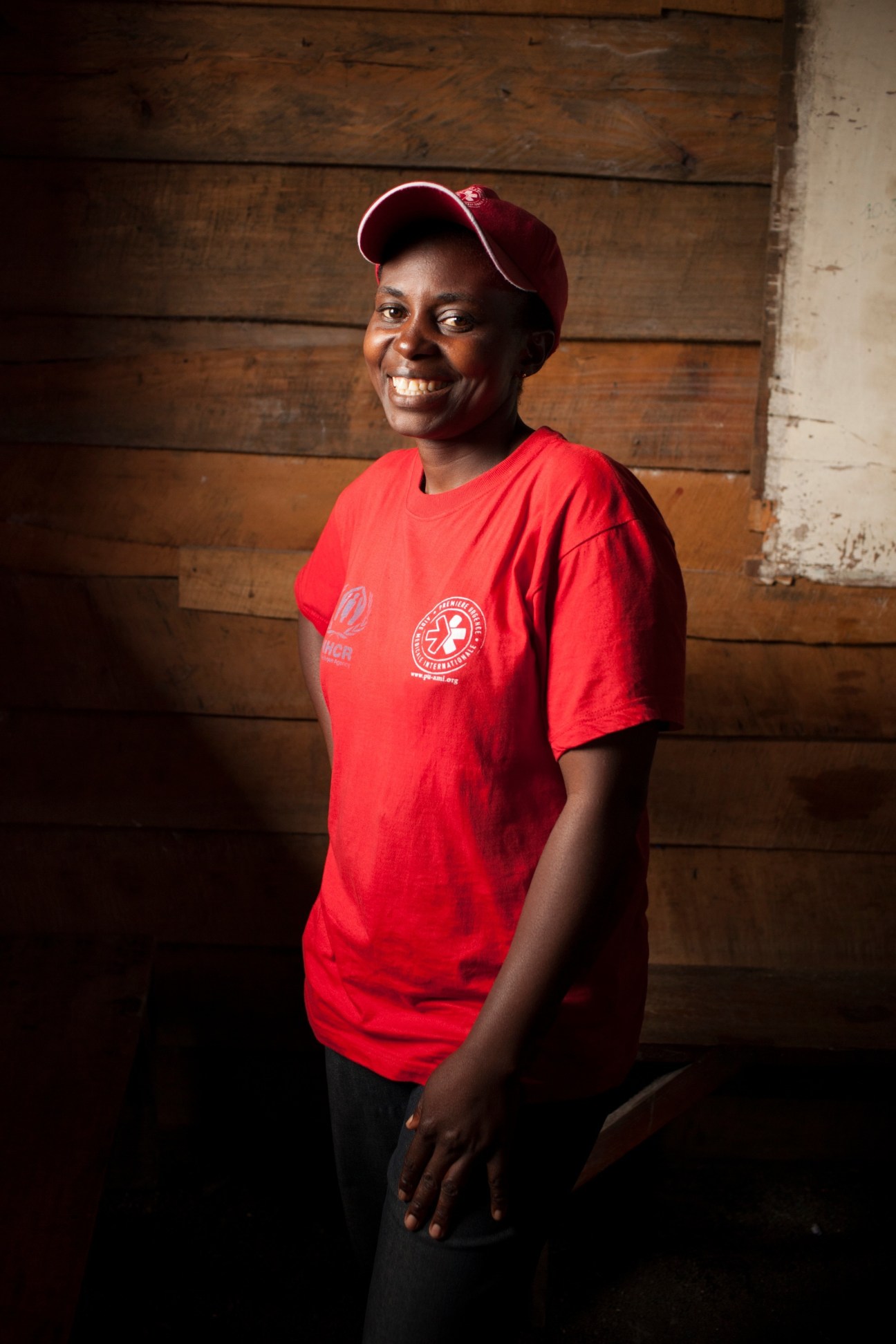 Woman in red Première Urgence aid organization t-shirt and maroon cap smiling at camera against wooden wall.