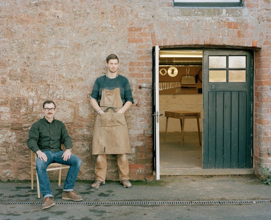 Two craftsmen pose outside a brick building with a green door; one seated, one standing in work apron.