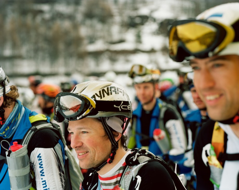 Military ski mountaineers in racing gear with helmets and backpacks prepare for competition.