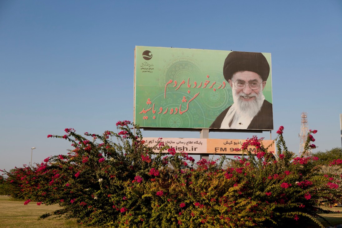 Billboard featuring Ayatollah Ruhollah Khomeini with Persian text against a green background, surrounded by pink flowering bushes.
