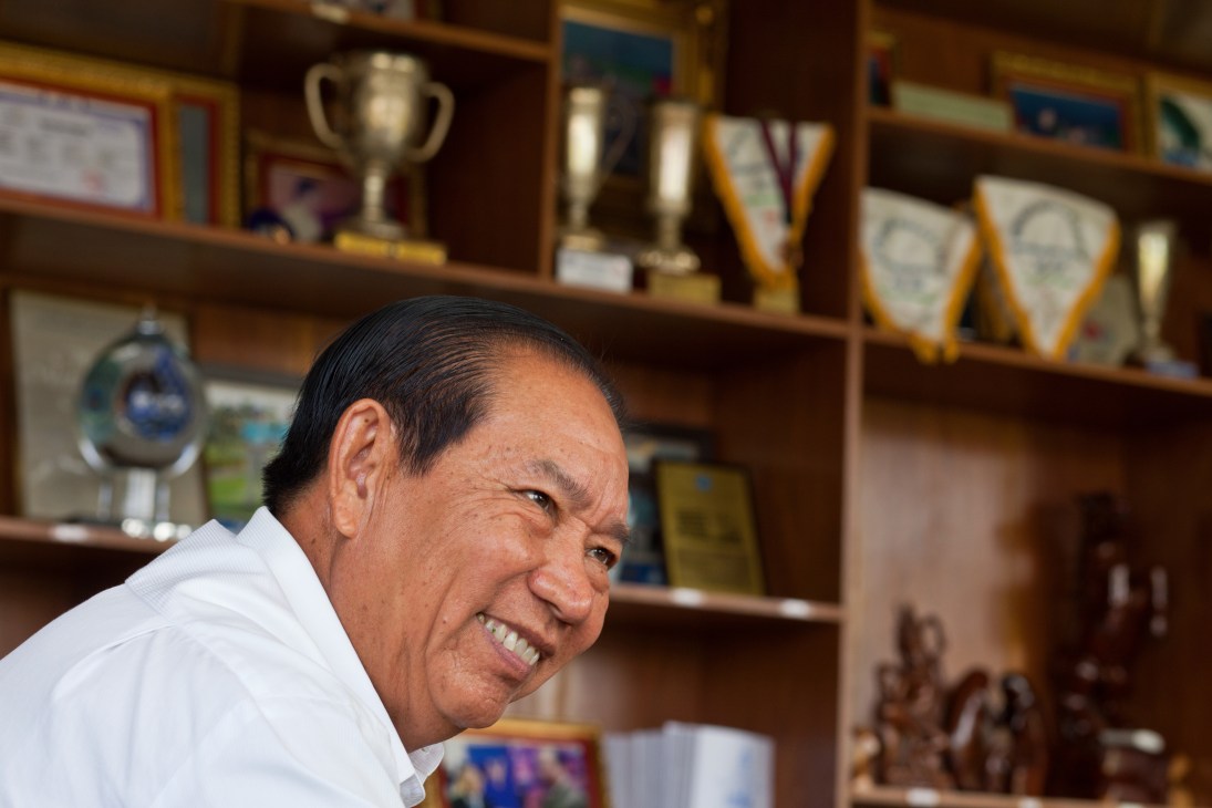 Man in white shirt smiling at camera with trophy and awards on shelf behind him.