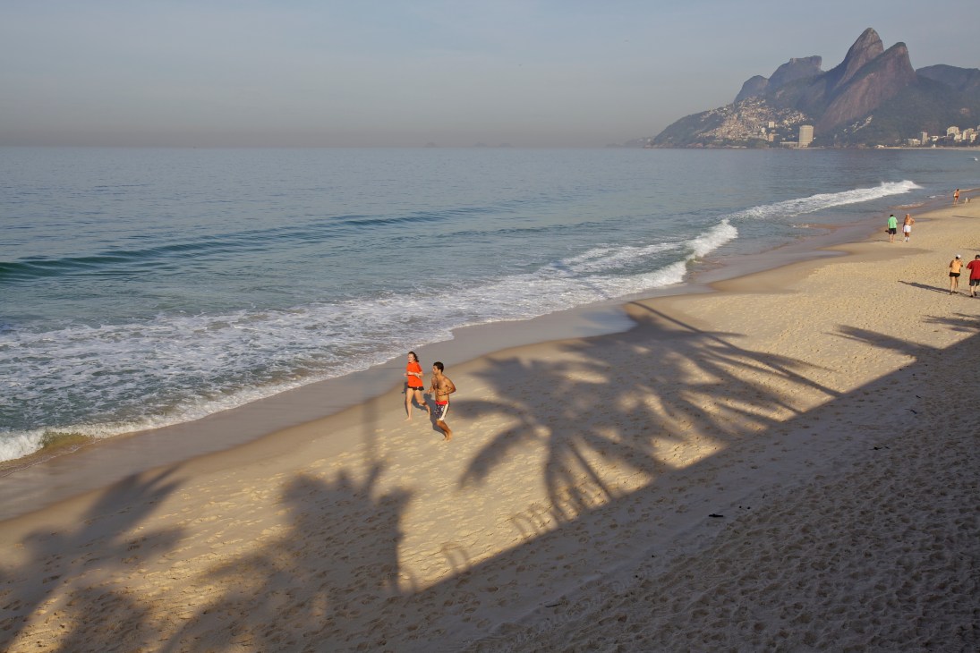 Joggers running on Rio de Janeiro's Ipanema Beach with mountains visible across the bay.