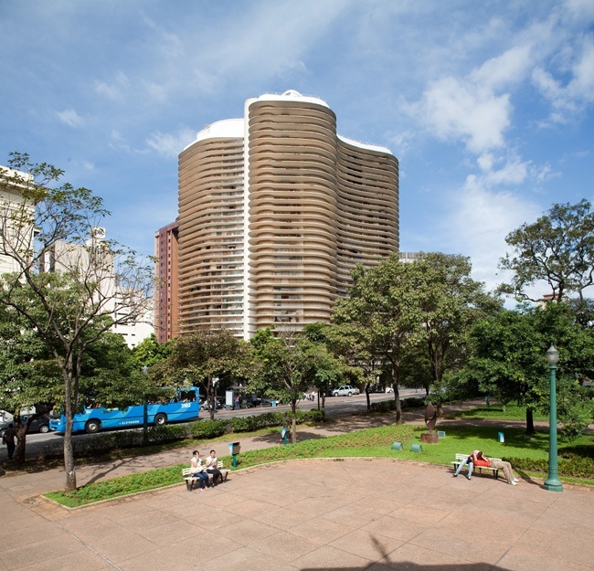 Curved modern residential tower with horizontal ribbed facade overlooks a public square with trees and a blue bus.