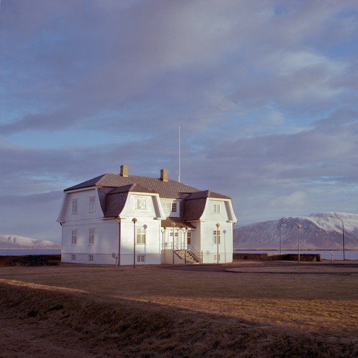 A white wooden house with mansard roof stands isolated on barren Icelandic coastland with mountains in the distance.