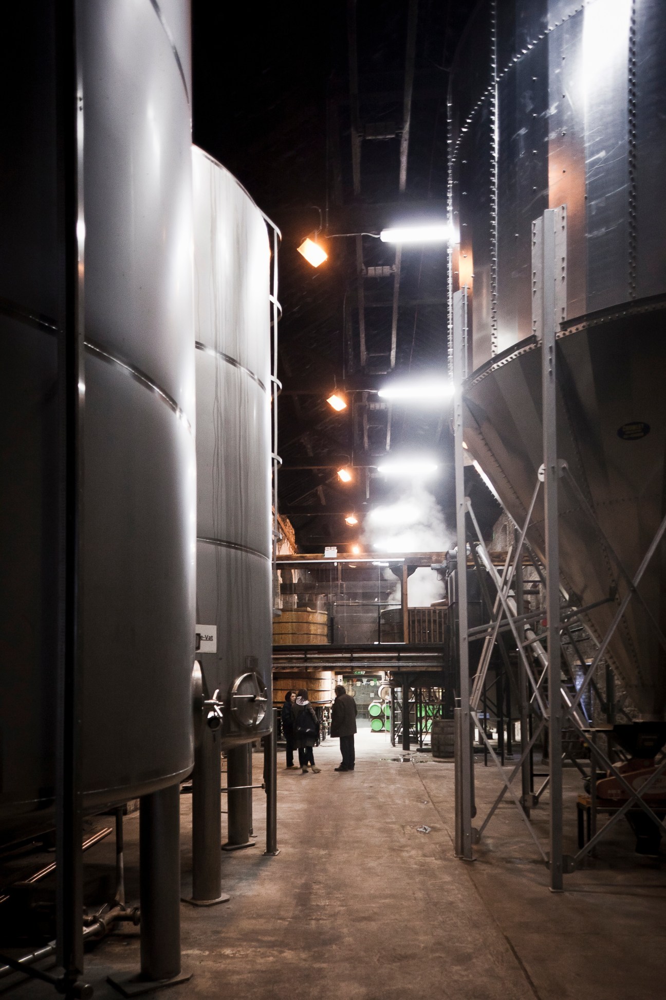 Interior of Cooley Distillery at night with large metal fermentation tanks and industrial equipment illuminated by overhead lighting.