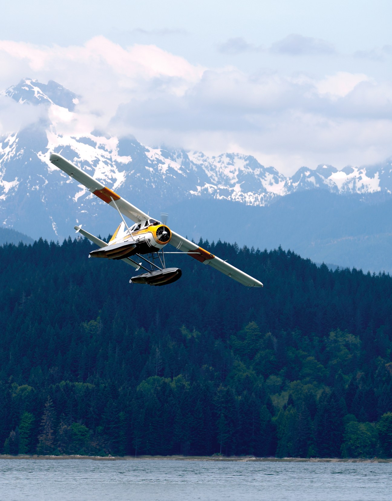 A Kenmore Air seaplane flies over forested islands with snow-capped mountains in the background.