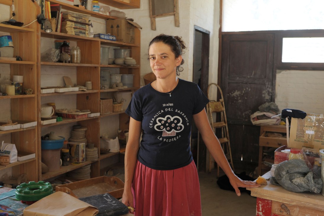 Serrana Dos Santos stands in her pottery workshop surrounded by wooden shelves holding ceramic pieces.