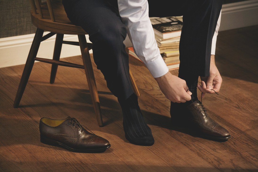 Man putting on brown leather oxfords with dark socks and trousers in an interior setting.