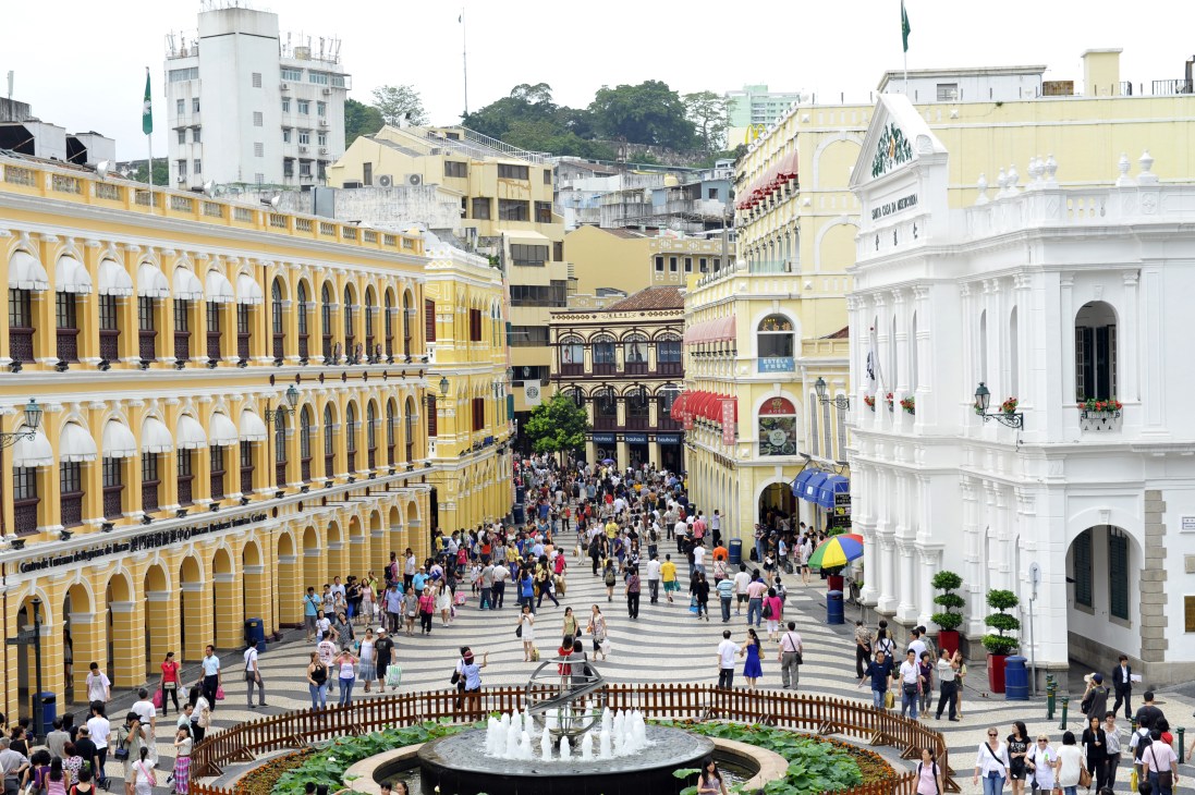 A bustling colonial square in Macau with colorful Portuguese architecture, traditional black and white patterned paving, and crowds of visitors.