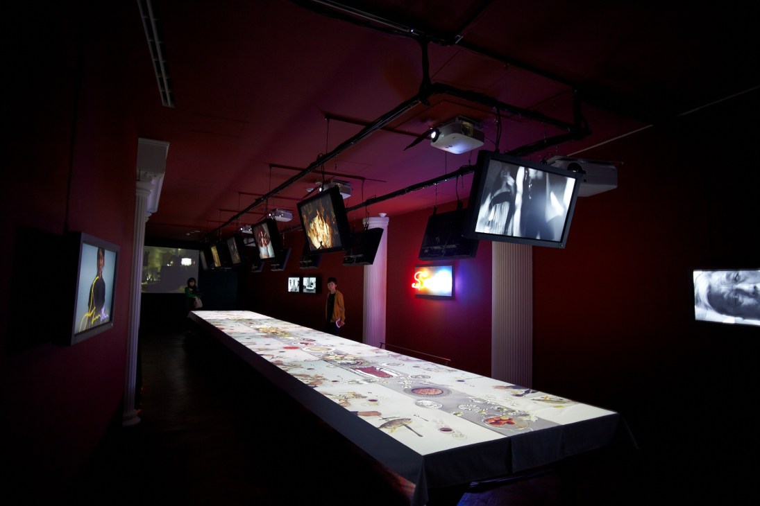 Contemporary art installation with illuminated display table and suspended video screens in magenta-walled gallery space.