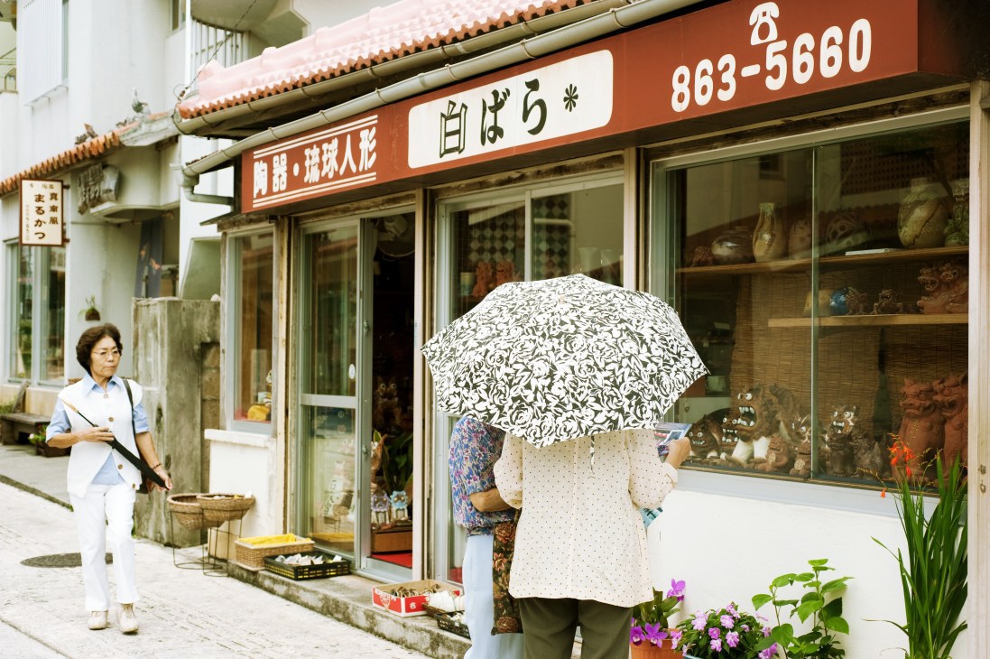 A street scene in Tsuboya with a woman holding a patterned umbrella standing in front of a traditional shop with red-tiled roof and Japanese signage.