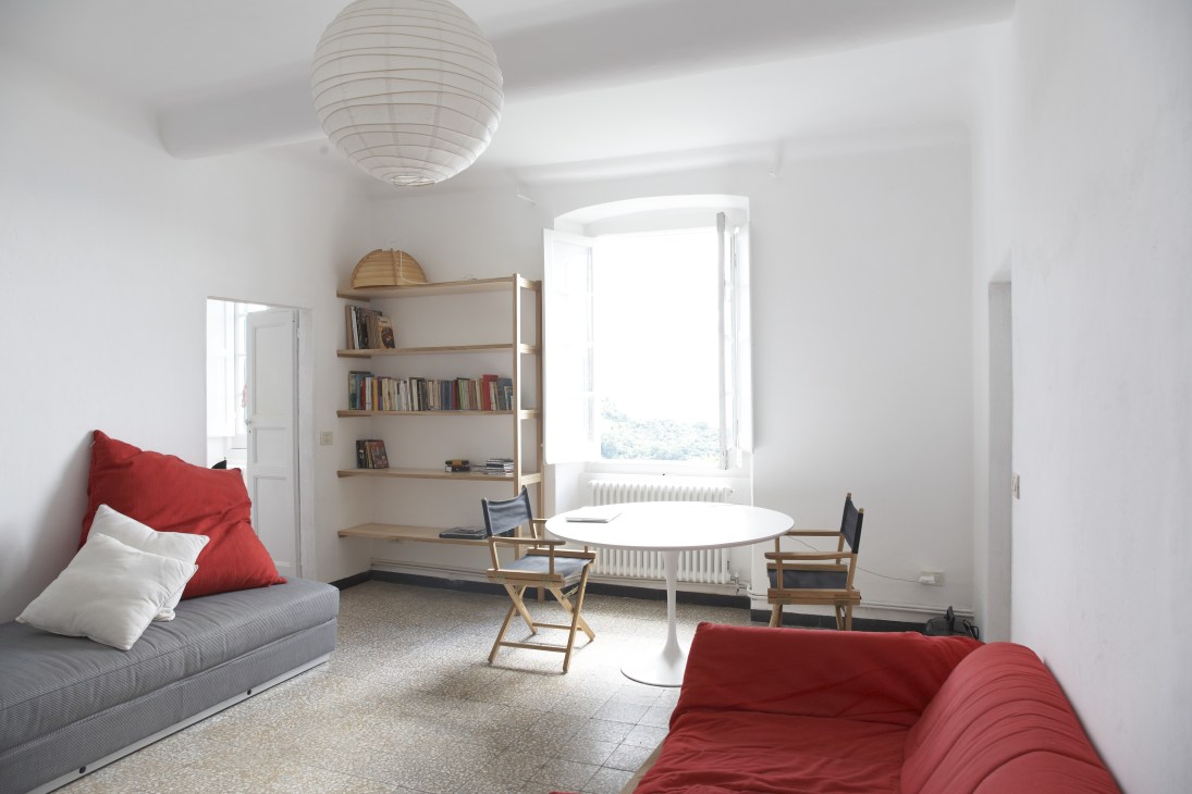 Minimalist living room with white walls, red sofas, wooden shelving, and bright windows overlooking landscape.