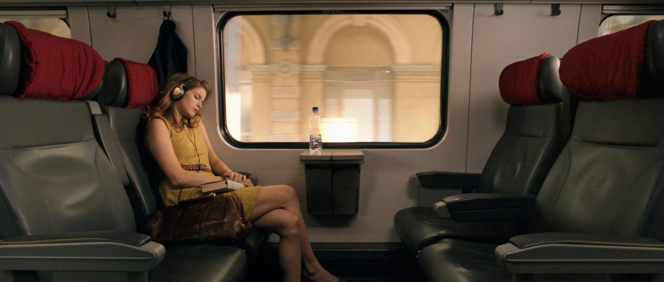 Woman sitting alone on a train car with red headrests and a window showing passing landscape.