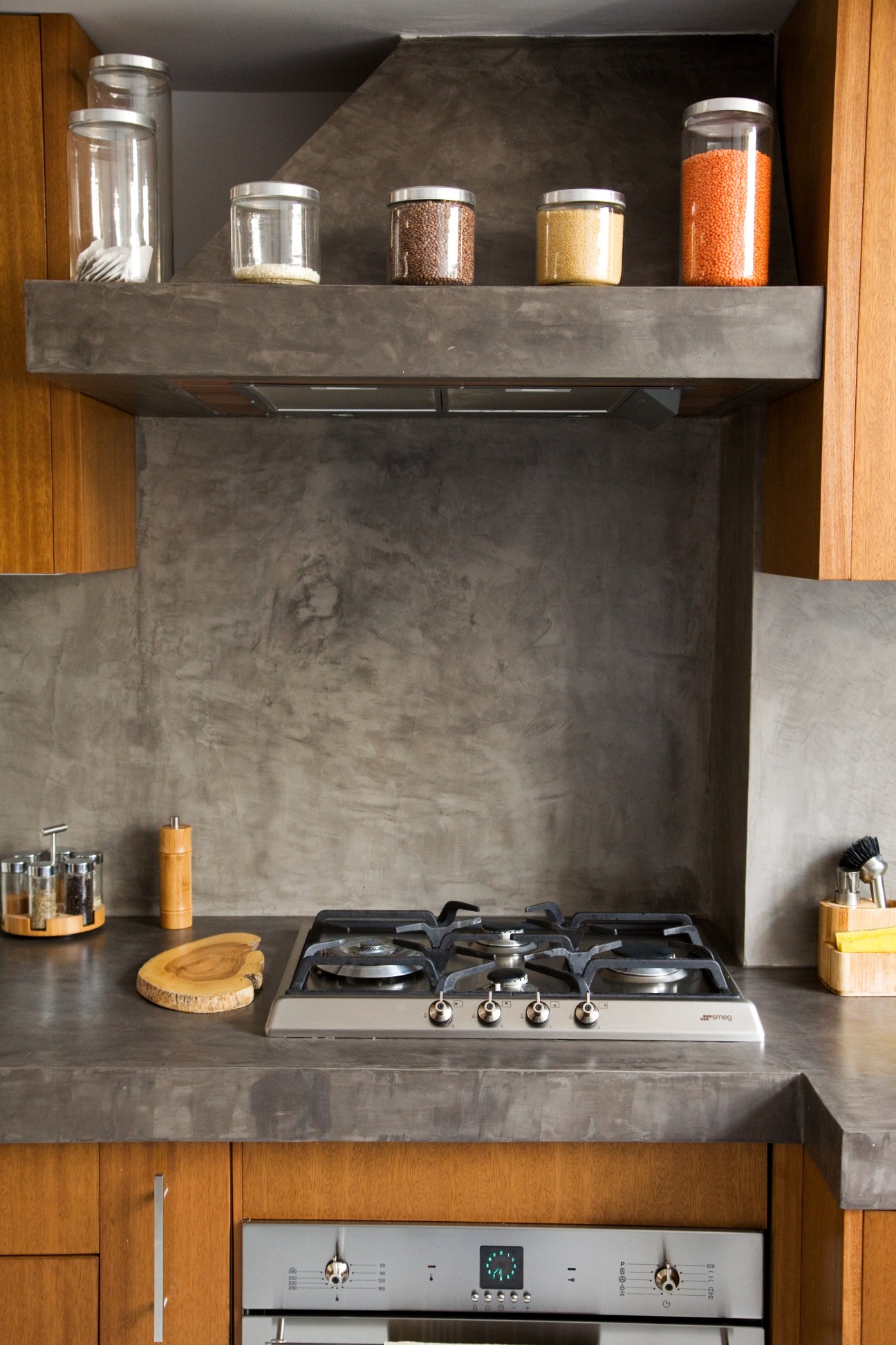 Modern kitchen with concrete backsplash, gas stovetop, wooden cabinetry, and shelf with glass storage jars.