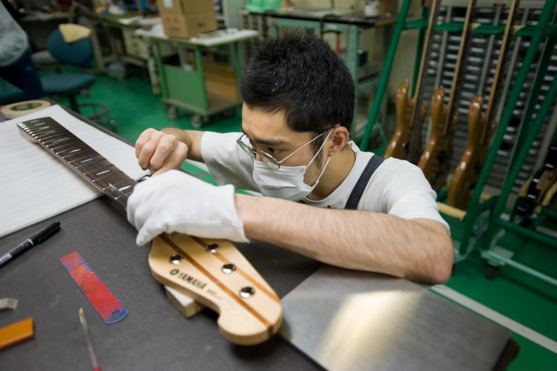 Craftsman wearing white gloves and mask assembles a guitar fingerboard in a factory assembly room.