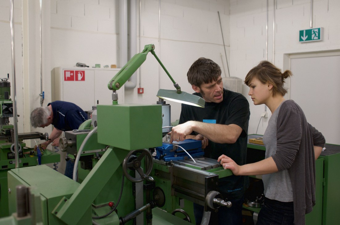 Workshop assistant instructs a student on operating green industrial machinery in a design studio.