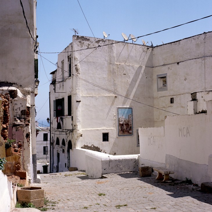 Deteriorating whitewashed buildings with peeling plaster line a narrow casbah street under clear blue sky.