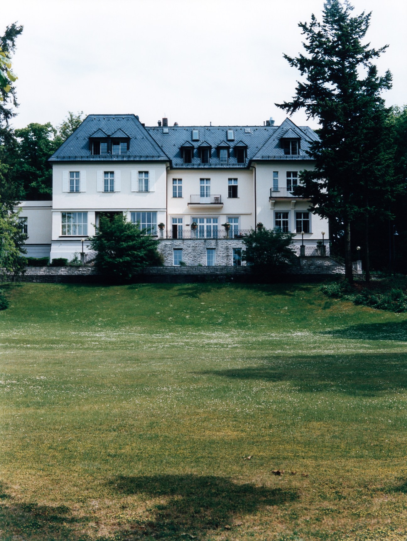 A grand Edwardian villa with white facade and mansard roof set on manicured grounds in Berlin's Wannsee district.