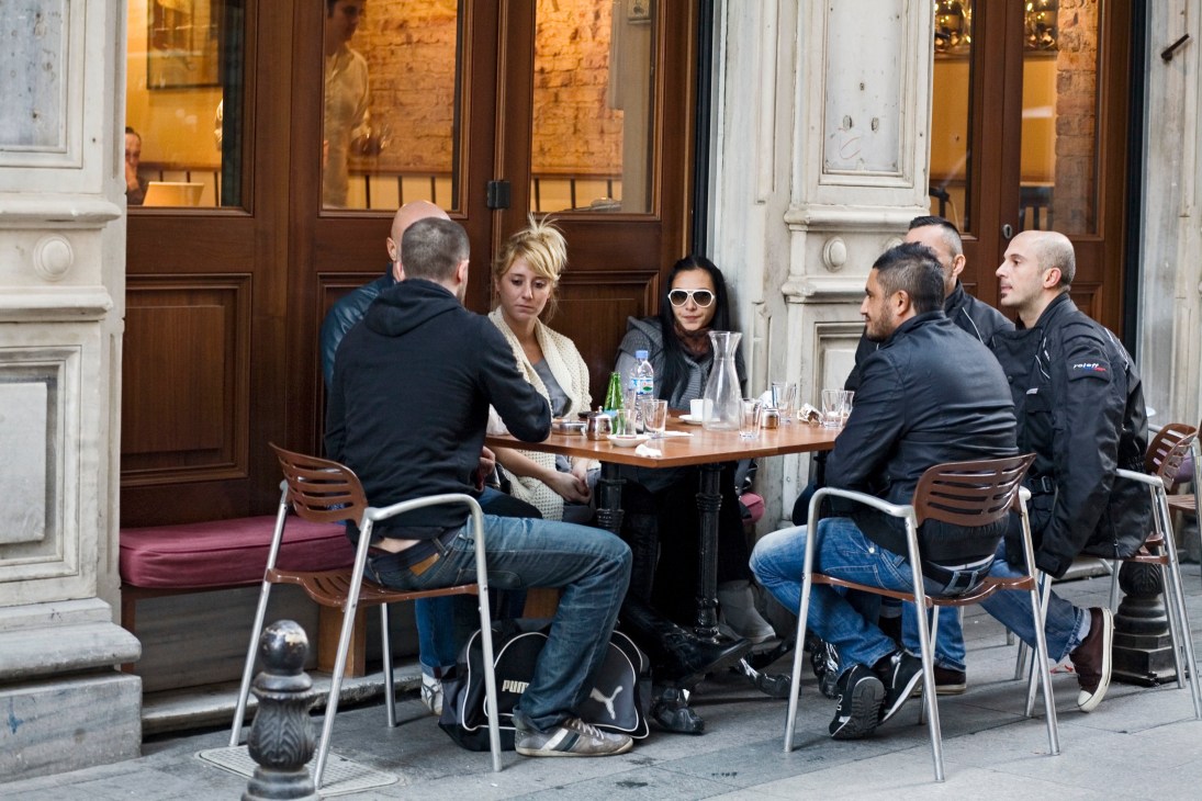 Group of people seated at outdoor café table in front of historic building with wooden doors.