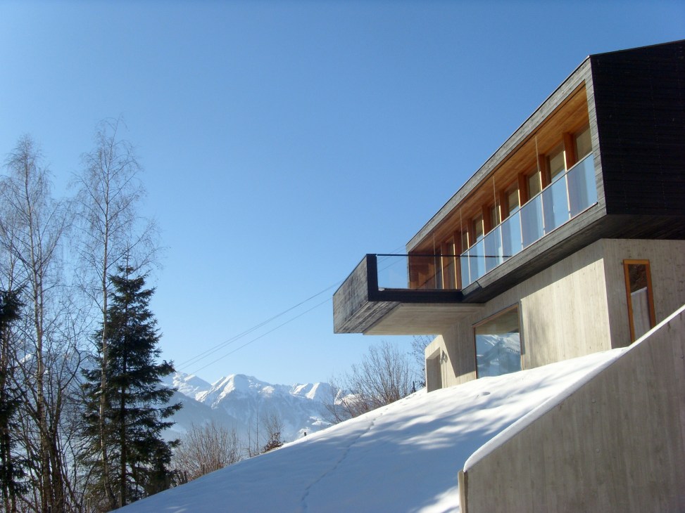 Modern mountain lodge with concrete and wood exterior overlooking snow-covered Alpine landscape.