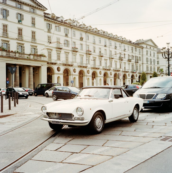 White vintage Fiat convertible parked in a Turin plaza with classical arcaded buildings behind.