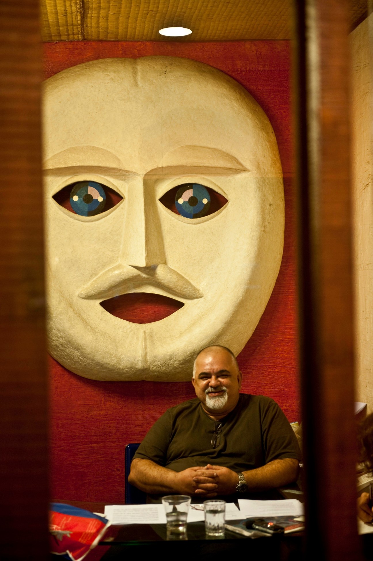Nikhil Kapoor sits at a desk beneath a large carved wooden mask with blue eyes and red mouth.