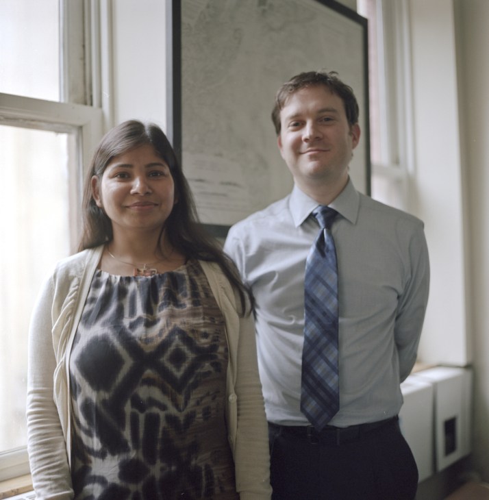 Two professionals standing in an office with windows behind them: a woman in a patterned dress and cardigan, and a man in a light blue shirt and dark tie.