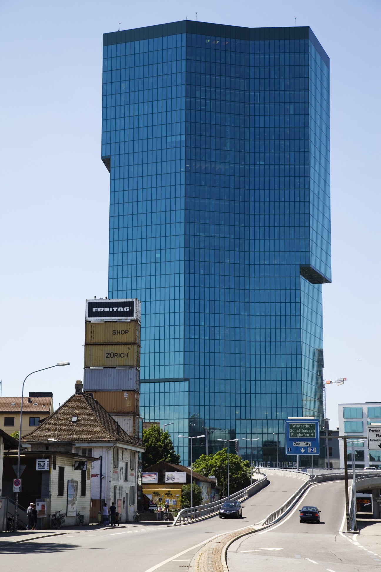 Modern blue-glass skyscraper next to traditional Swiss building and Freitag shipping container shop in Zurich.