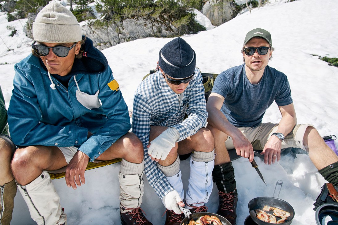 Three men sit in snow eating from pans during a high-altitude lunch break in the mountains.
