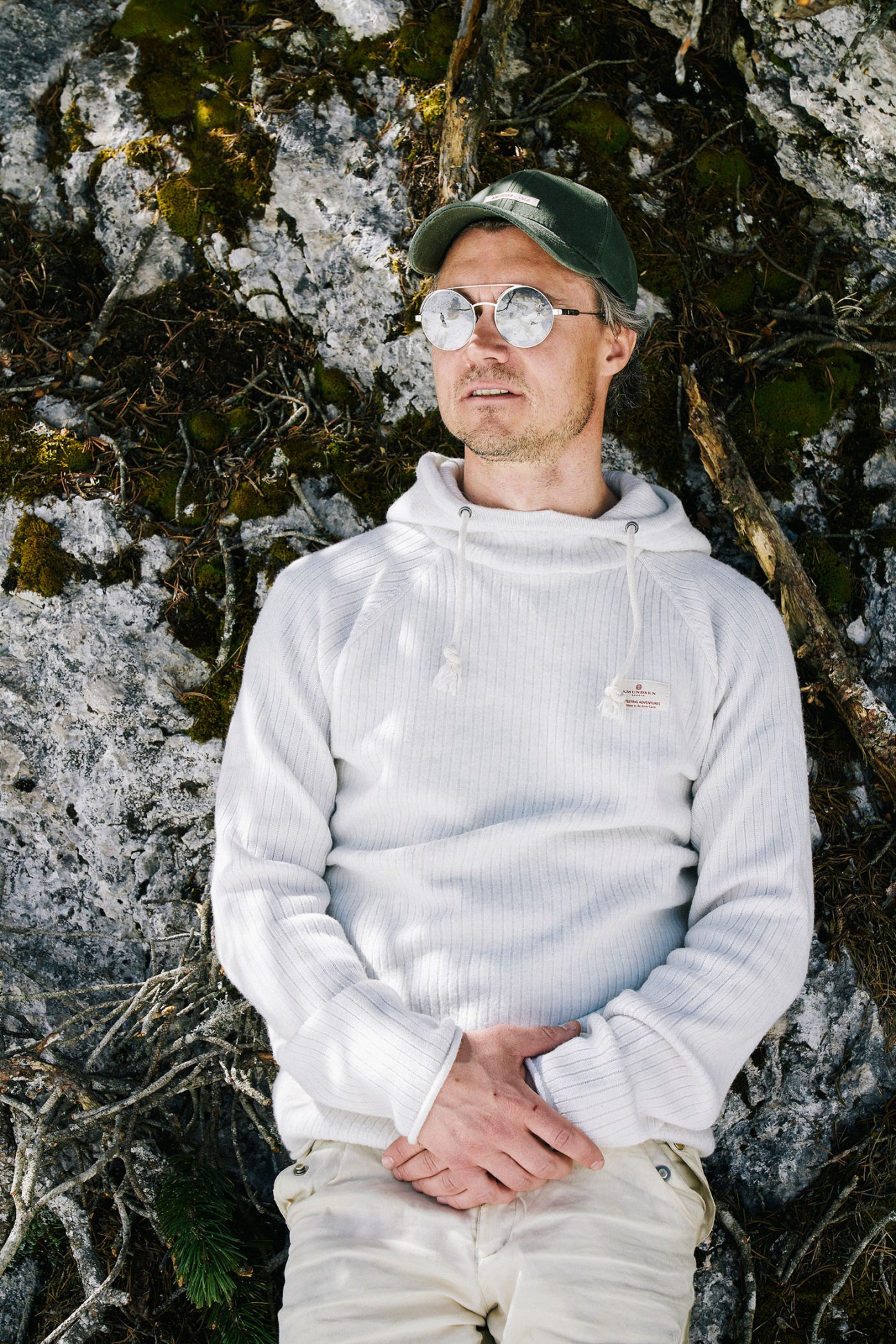 Man in white boiled-wool hoodie and waxed-cotton cap leaning against moss and lichen-covered rock.