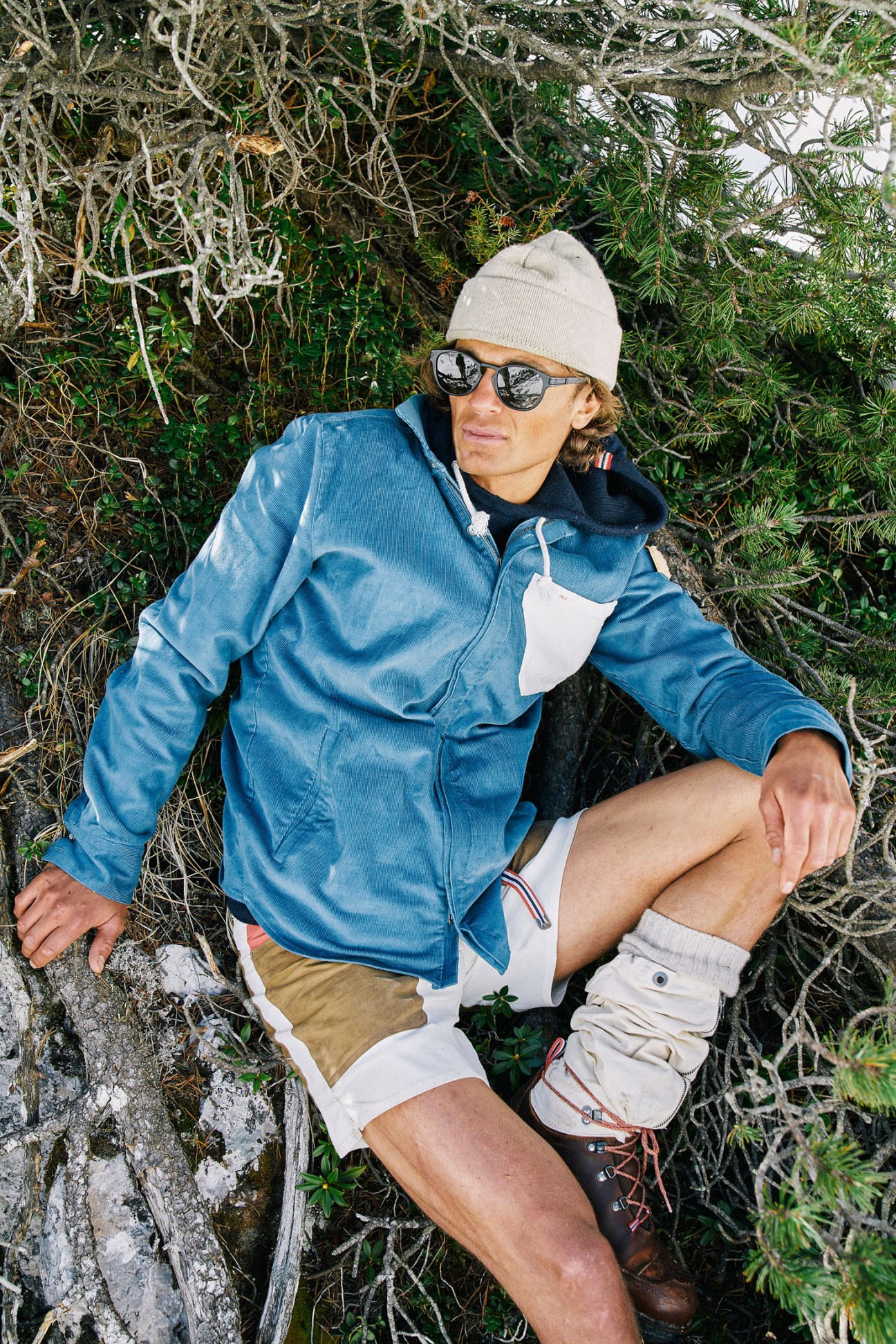 Man wearing Amundsen Sports denim jacket and white shorts crouches against vegetation during mountain hiking expedition.