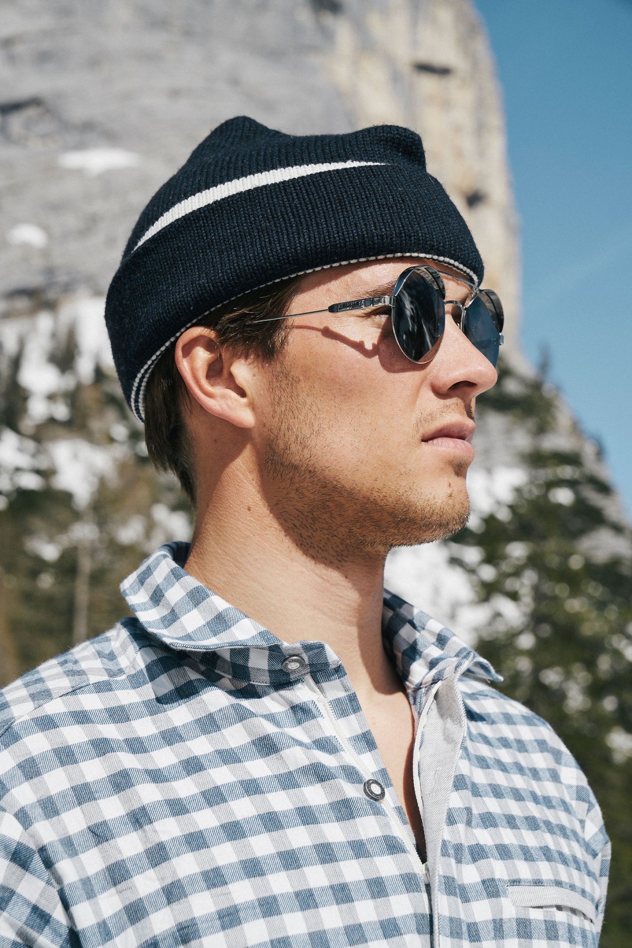Man wearing black beanie with white stripe and round sunglasses in snowy mountain landscape