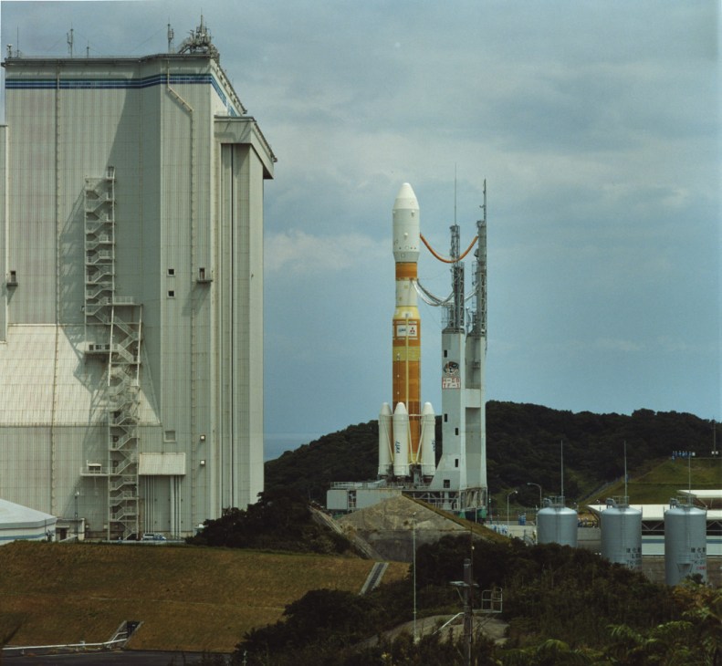 Japanese rocket with white nose cone and brown fuel tank on launch pad next to service tower.