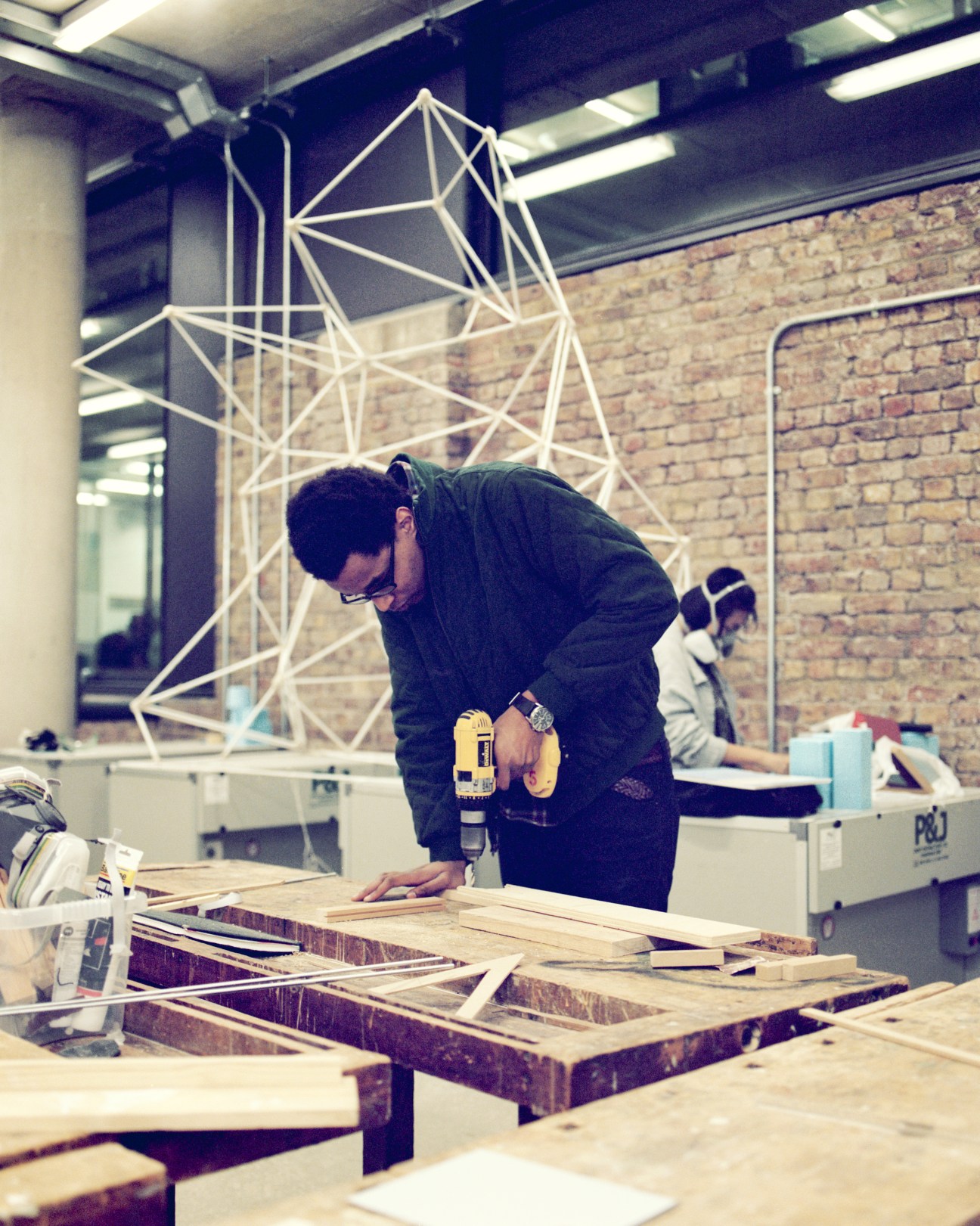Student working on wooden materials in a workshop with a geometric white sculpture in the background.