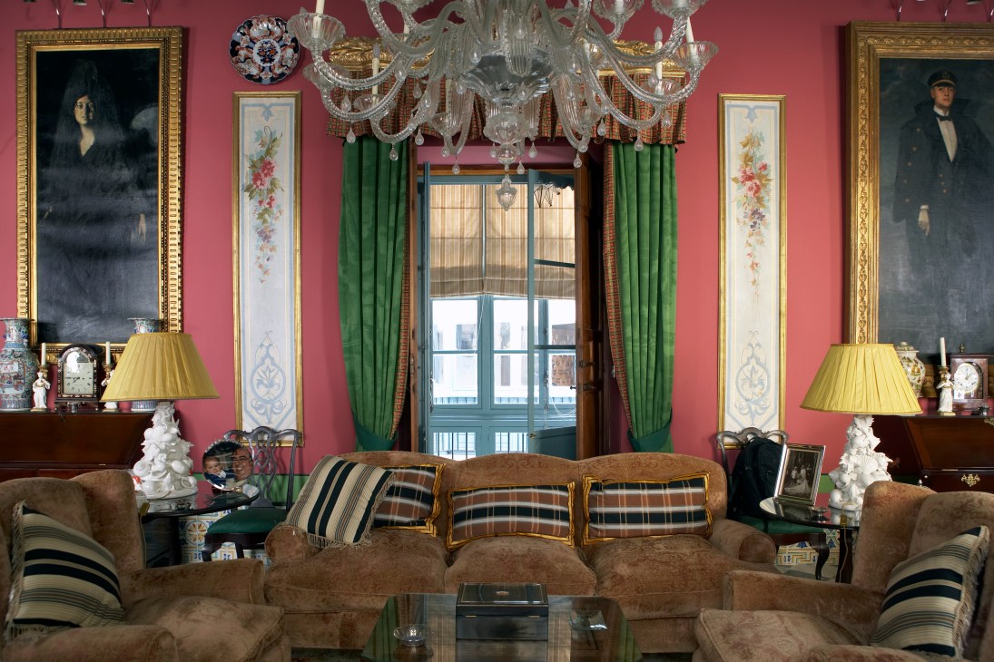Ornate interior of La Bella Escondida with pink walls, crystal chandelier, framed portraits, and brown upholstered seating.