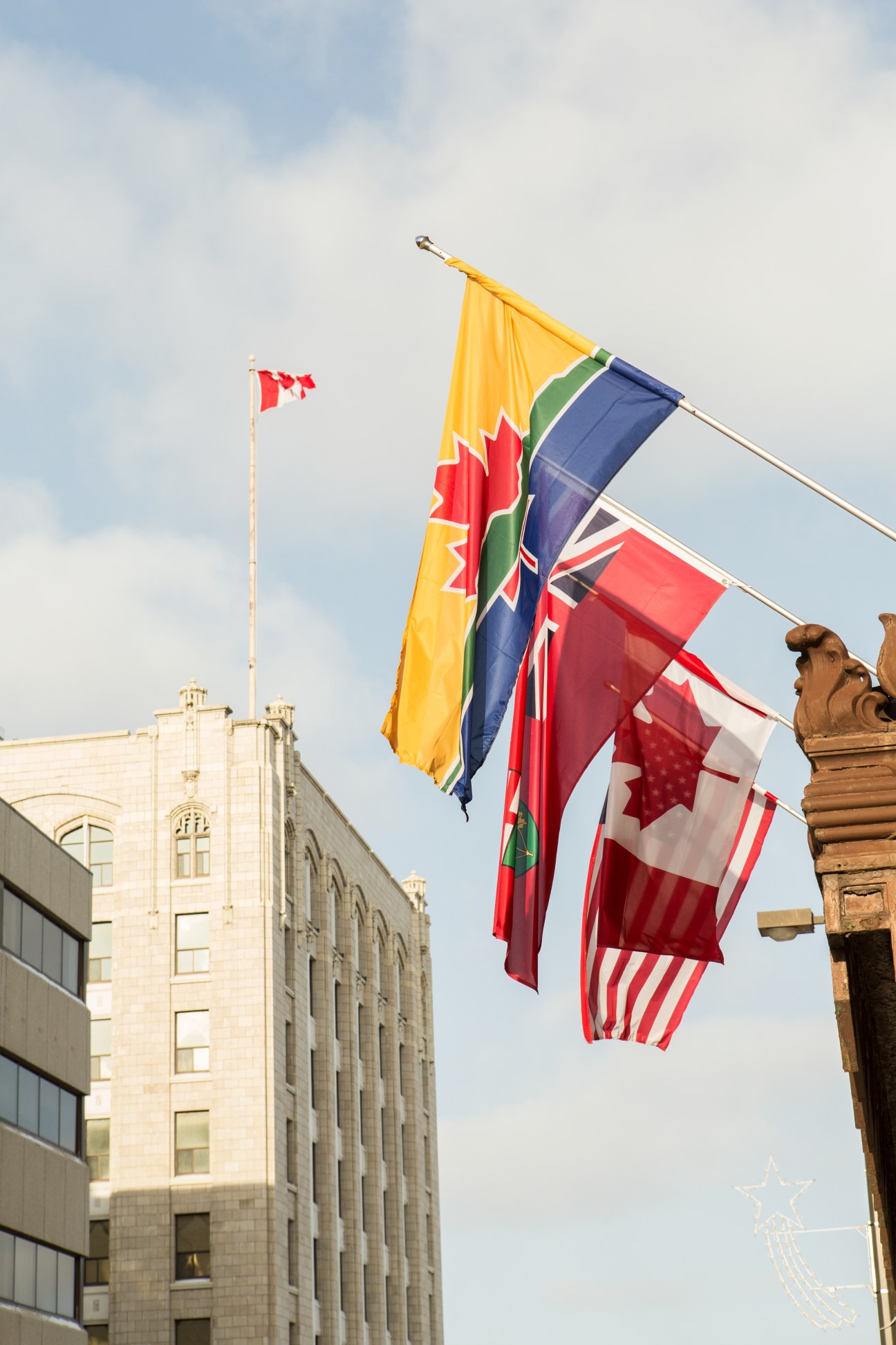 Multiple flags including Canadian and regional flags displayed against a stone building and blue sky.