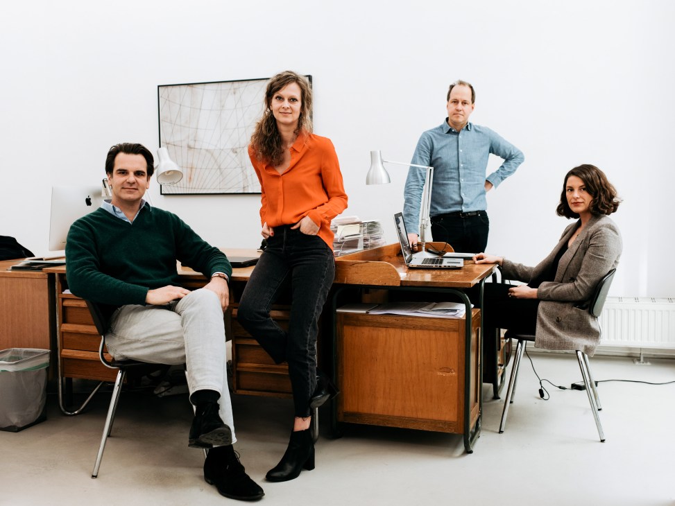 Four members of the Phileas team pose together in their Vienna office, with man seated left in green sweater, woman standing in orange shirt, man in blue shirt standing center, and woman seated right in brown blazer.