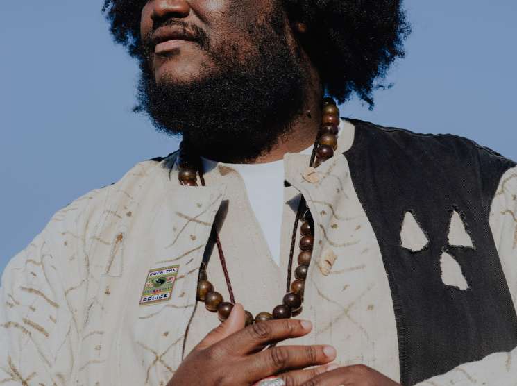 Kamasi Washington wears an African-inspired cream jacket with beaded necklace against a blue sky background.
