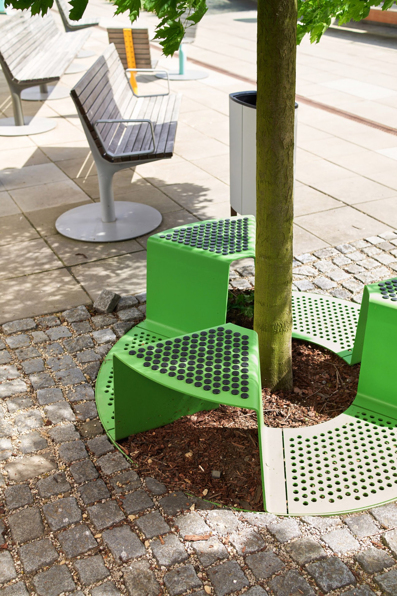 Green perforated tree guards and benches surrounding a tree trunk in a paved outdoor area.