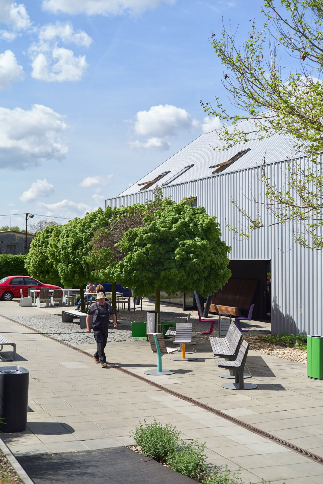 Mmcité's open-air showroom pathway lined with benches, bins and street furniture beneath a modern industrial building with trees.